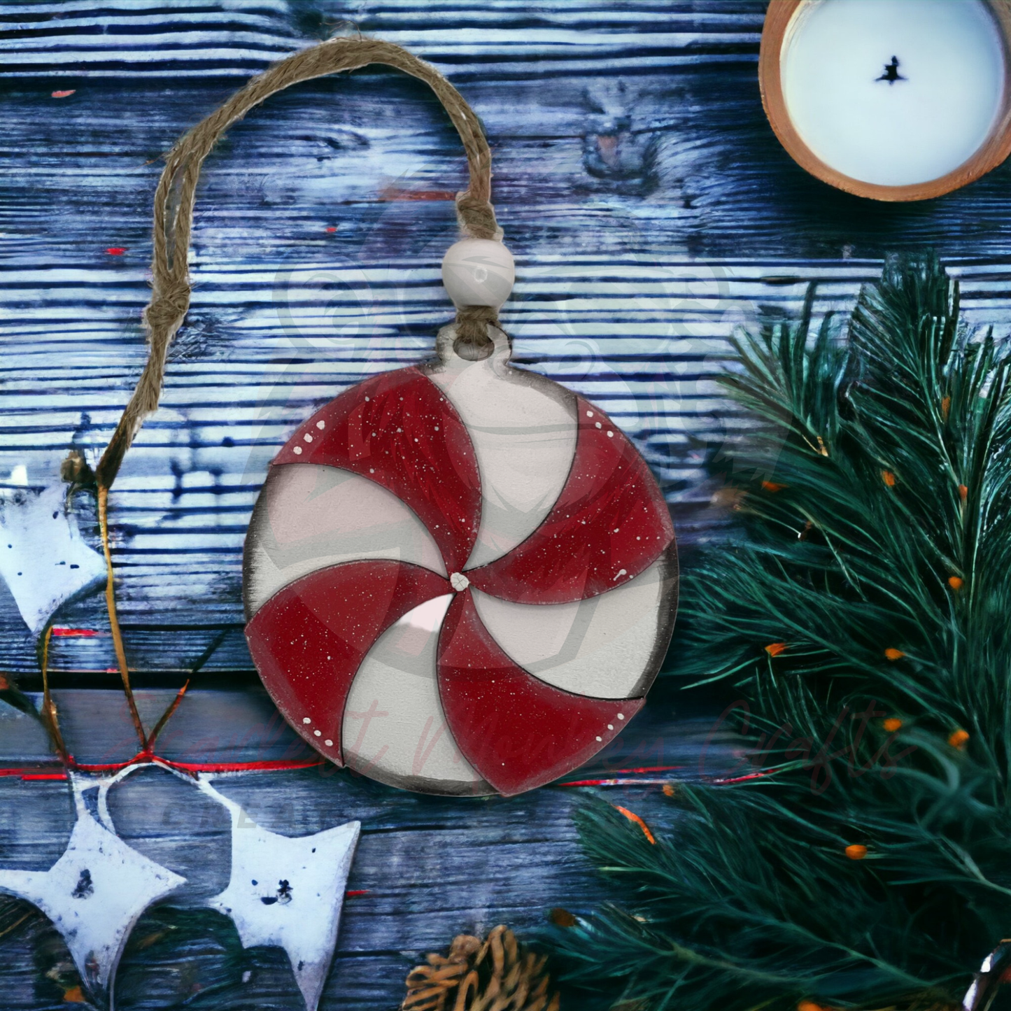 Hand-painted red and white pinwheel Christmas ornament with rustic twine and bead hanging loop on wooden background with greenery and candle