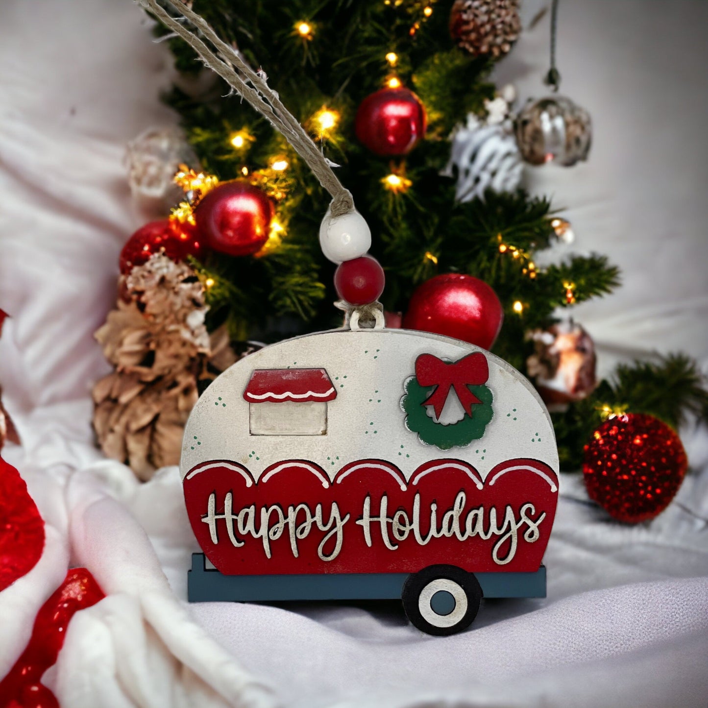 Handcrafted Christmas ornament with "Happy Holidays" camper design in front of festive tree.