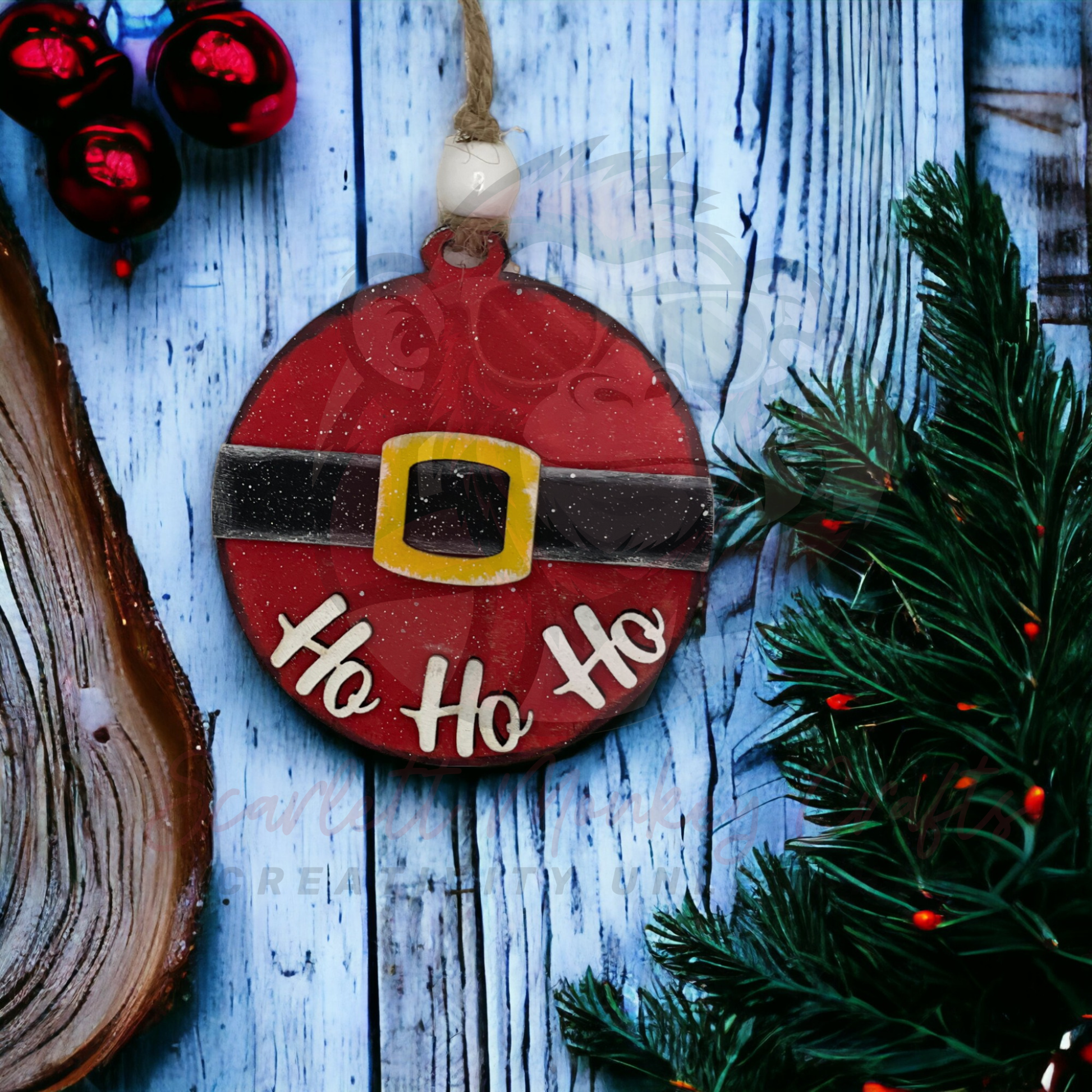 Hand-painted wooden Christmas ornament with Santa belt and Ho Ho Ho lettering hanging on twine against a blue wooden background and green pine branches