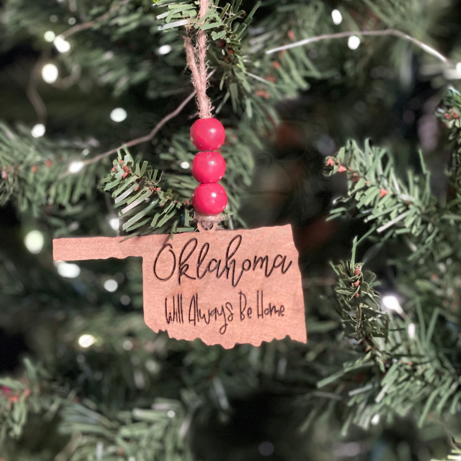 Hand-painted wooden Oklahoma-shaped Christmas ornament with "Oklahoma Will Always Be Home" lettering and red bead accents hanging on tree