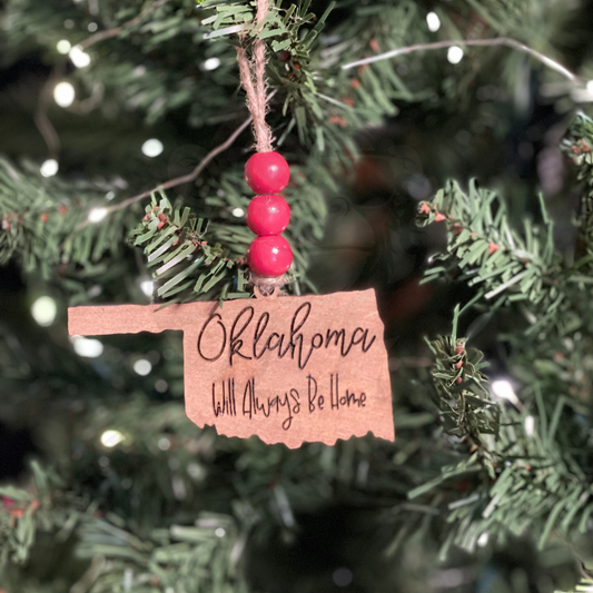 Hand-painted wooden Oklahoma-shaped Christmas ornament with "Oklahoma Will Always Be Home" lettering and red bead accents hanging on tree